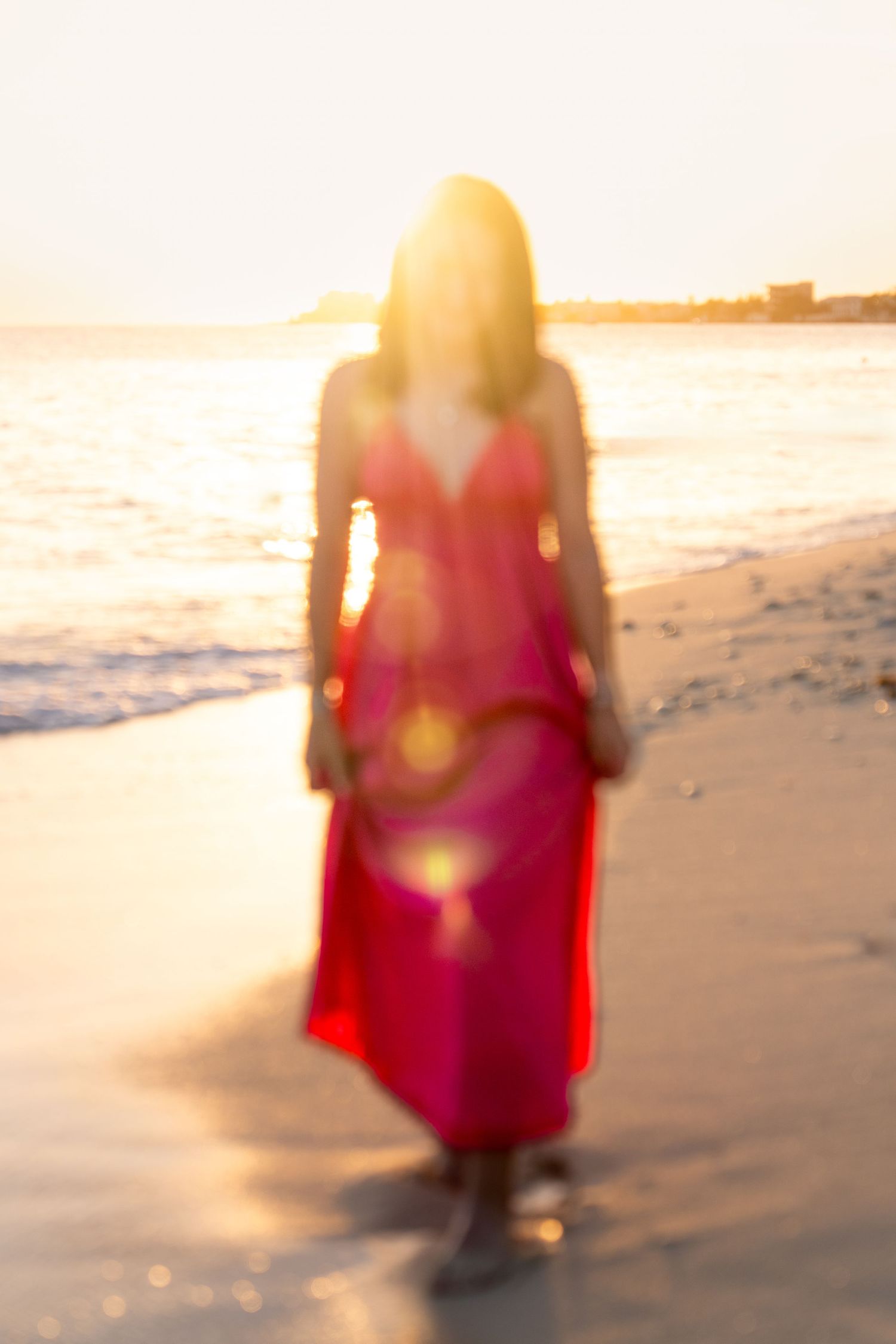 Dreamy silhouette of someone in a red dress walking along a sunlit beach at dusk.