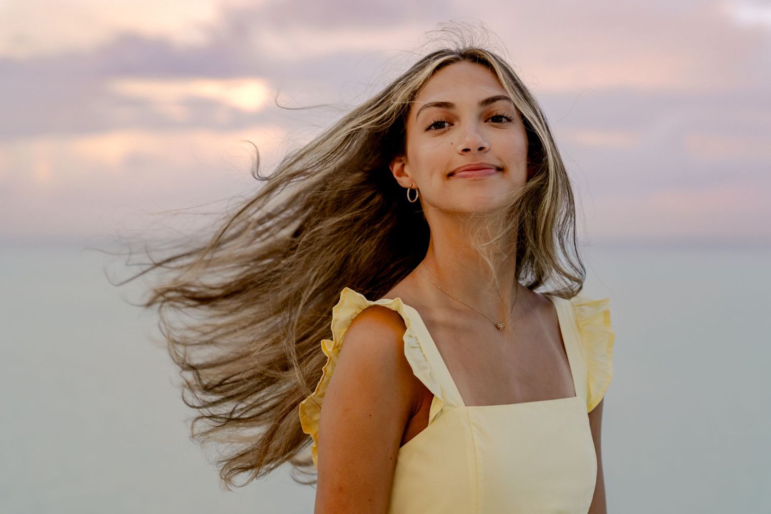 Long hair flows in the wind as someone in a yellow dress stands against a dusky beach backdrop.