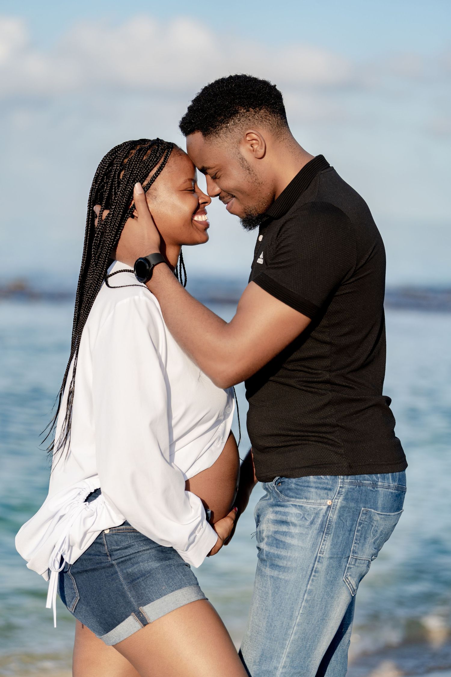 Romantic couple in casual attire shares intimate moment by ocean during sunset photoshoot.