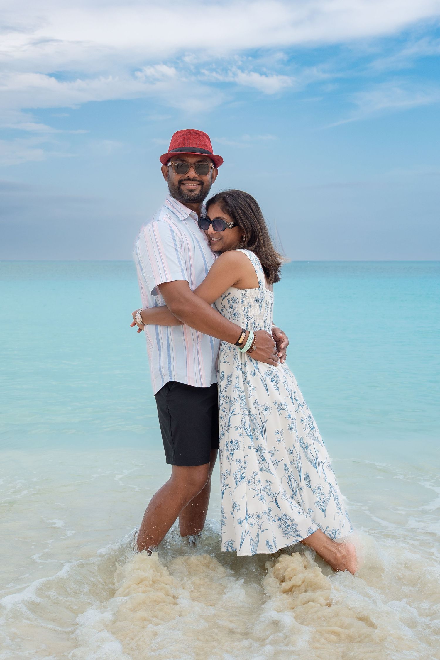 Couple embracing in turquoise ocean water, man in red hat and woman in floral dress, tropical beach vacation.