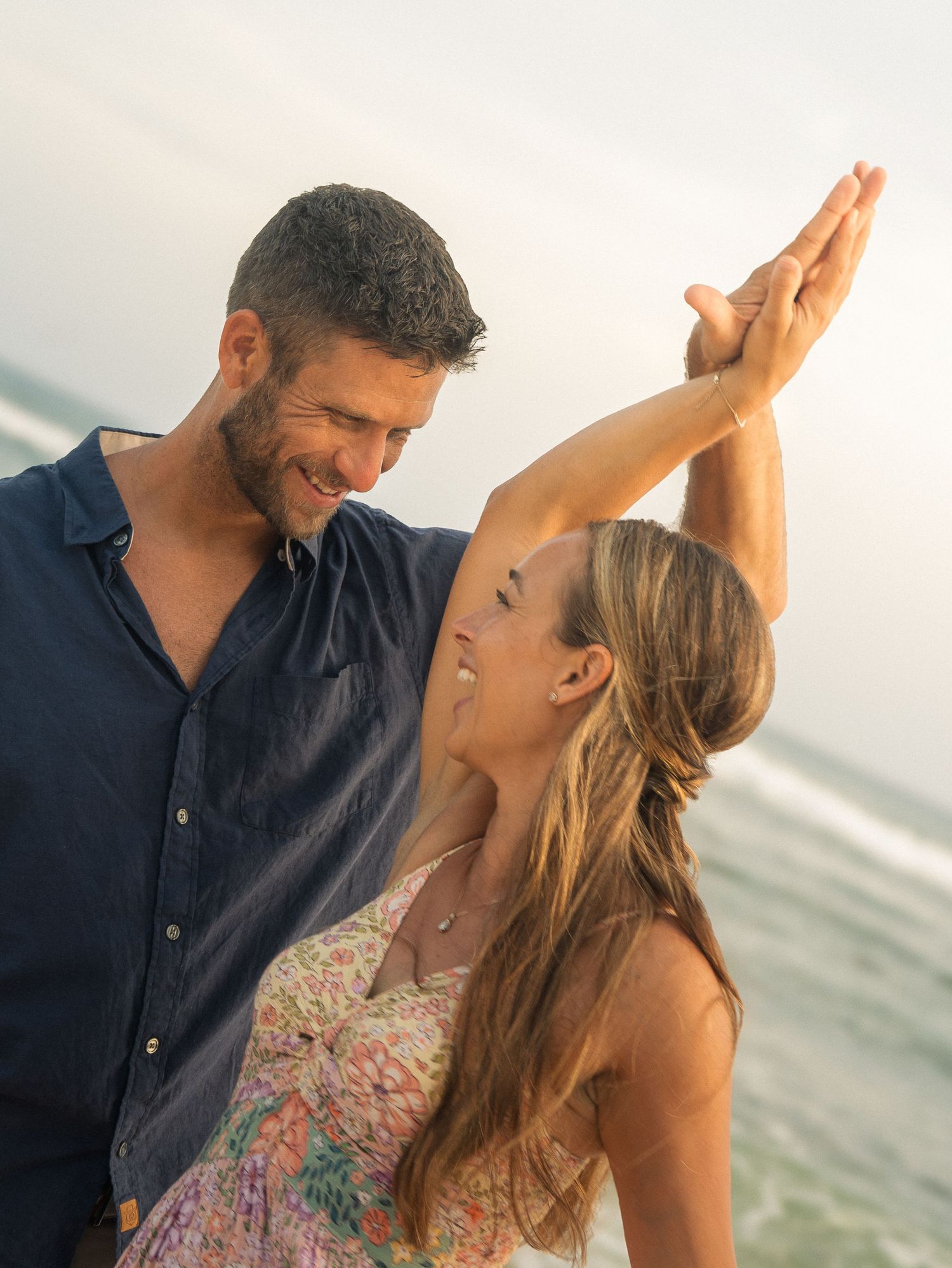 Couple dancing on a beach at sunset, man spinning woman in floral sundress by the ocean.