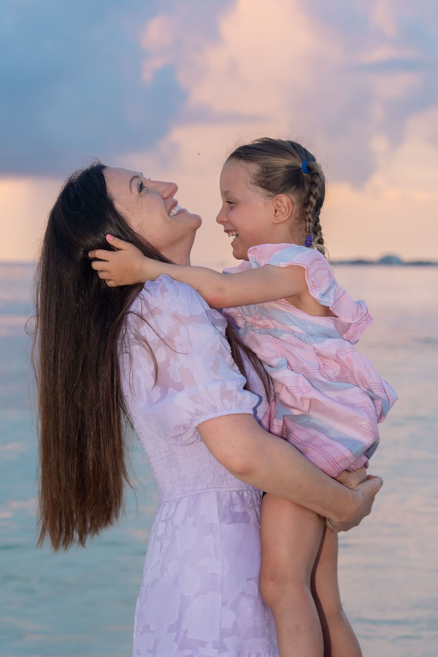 A mother holds her smiling daughter near a turquoise ocean at sunset, both wearing matching pink dresses.