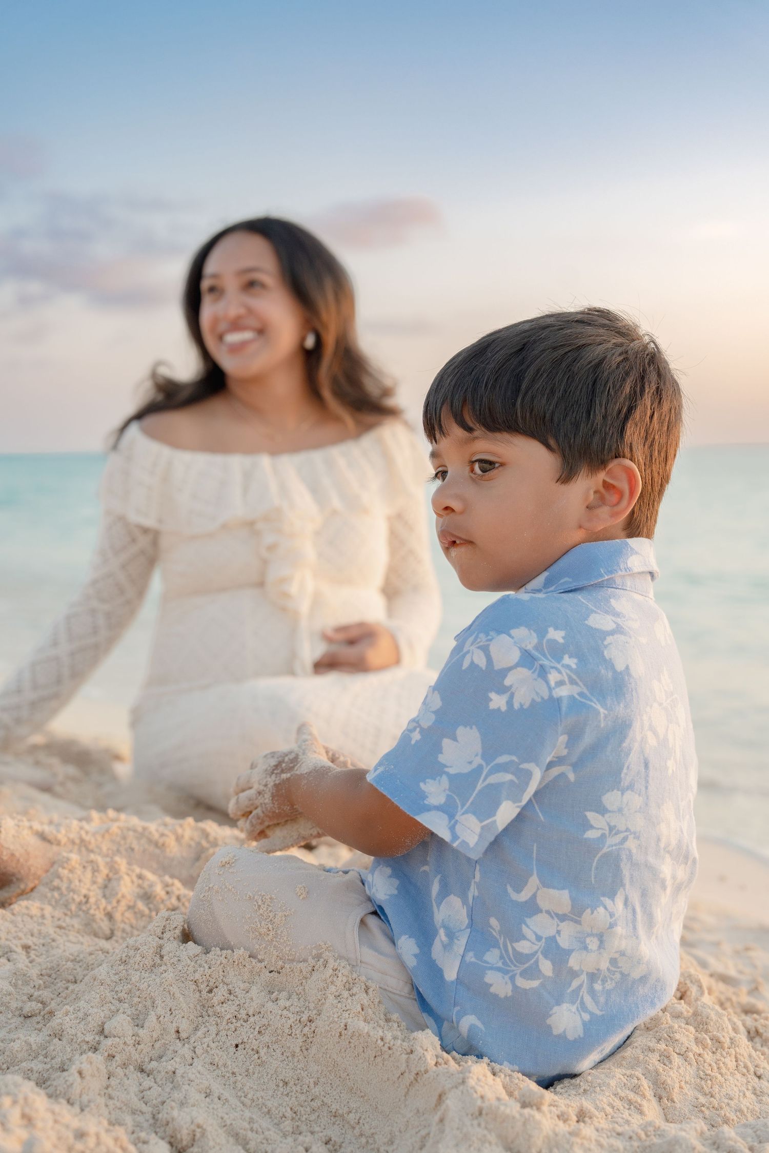 A young boy in a floral shirt plays in the sand on a tropical beach while a smiling woman sits behind him.