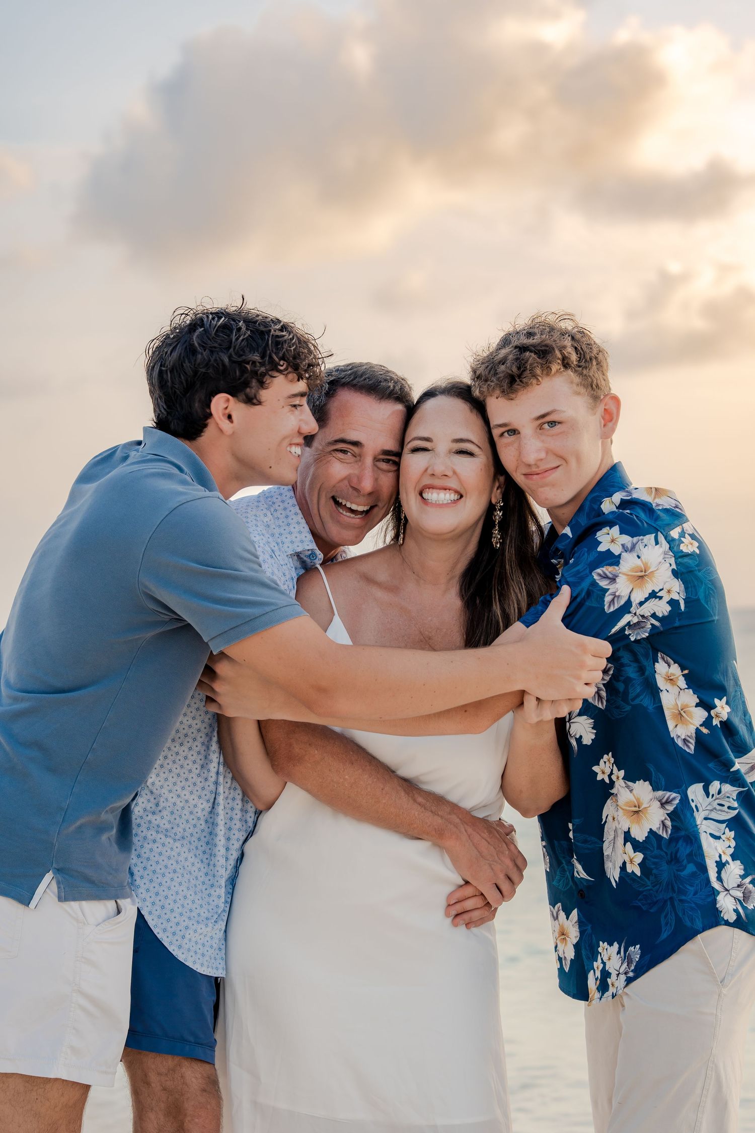 Happy family of four embracing on a beach at sunset, wearing blue and white outfits.