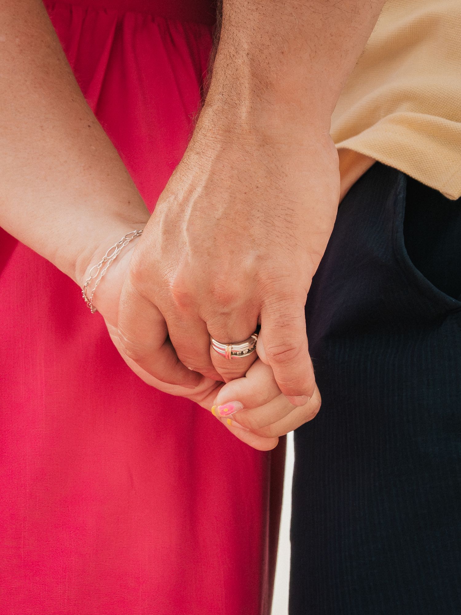Close-up of a couple holding hands, woman wearing a delicate bracelet and man with a silver ring, dressed in pink and navy.