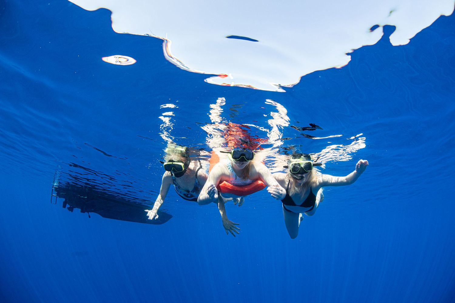 Snorkelers swim in deep blue ocean waters while boats float on the surface above.