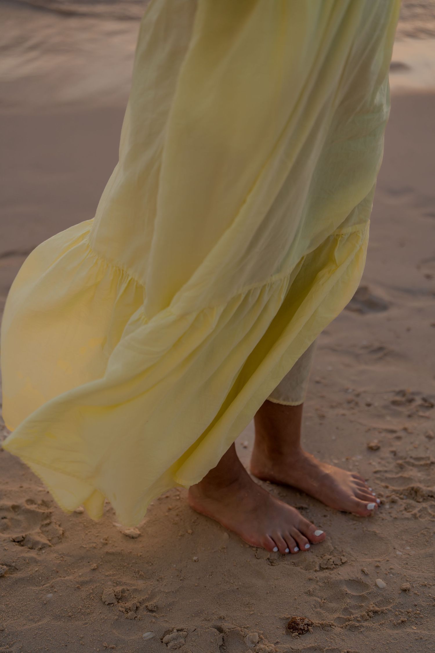Close-up of bare feet on sand with flowing yellow dress fabric in beach setting.