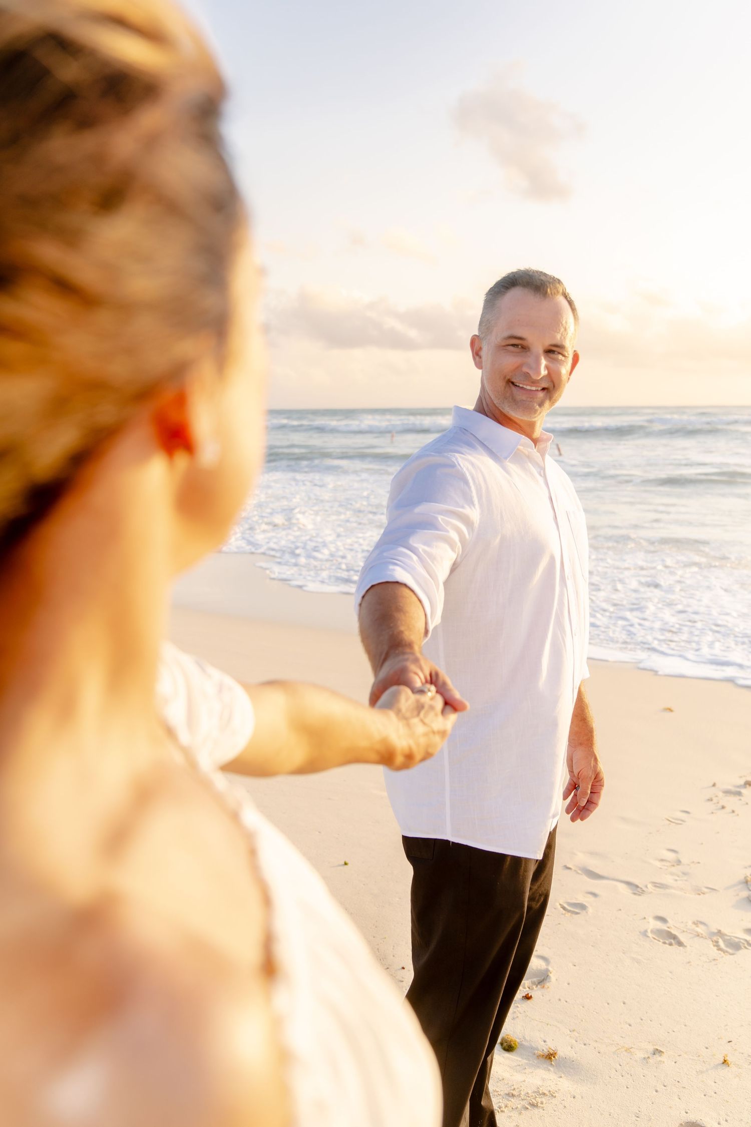 Couple in white clothing poses playfully on sandy beach during golden hour.