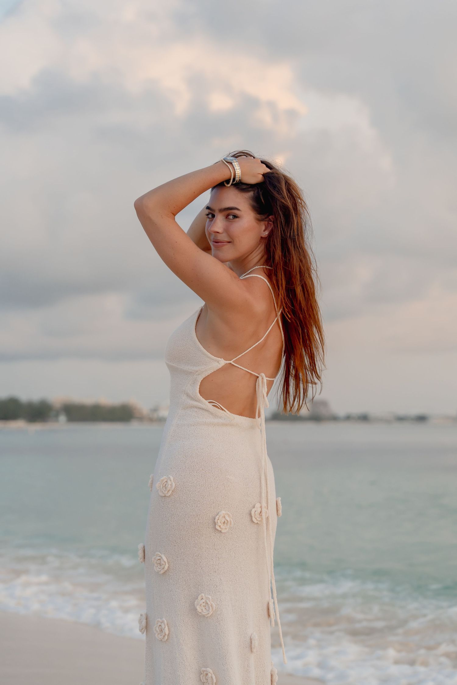 Young woman in white backless floral dress poses on a beach at sunset with ocean waves behind her.