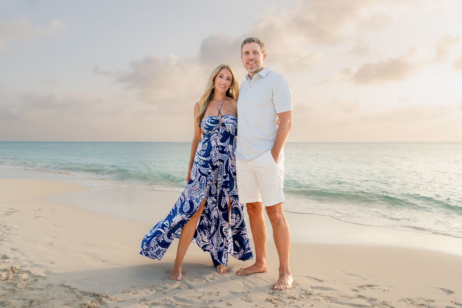 Couple posing on a sandy beach at sunset, woman in blue floral maxi dress, man in white shirt and shorts.