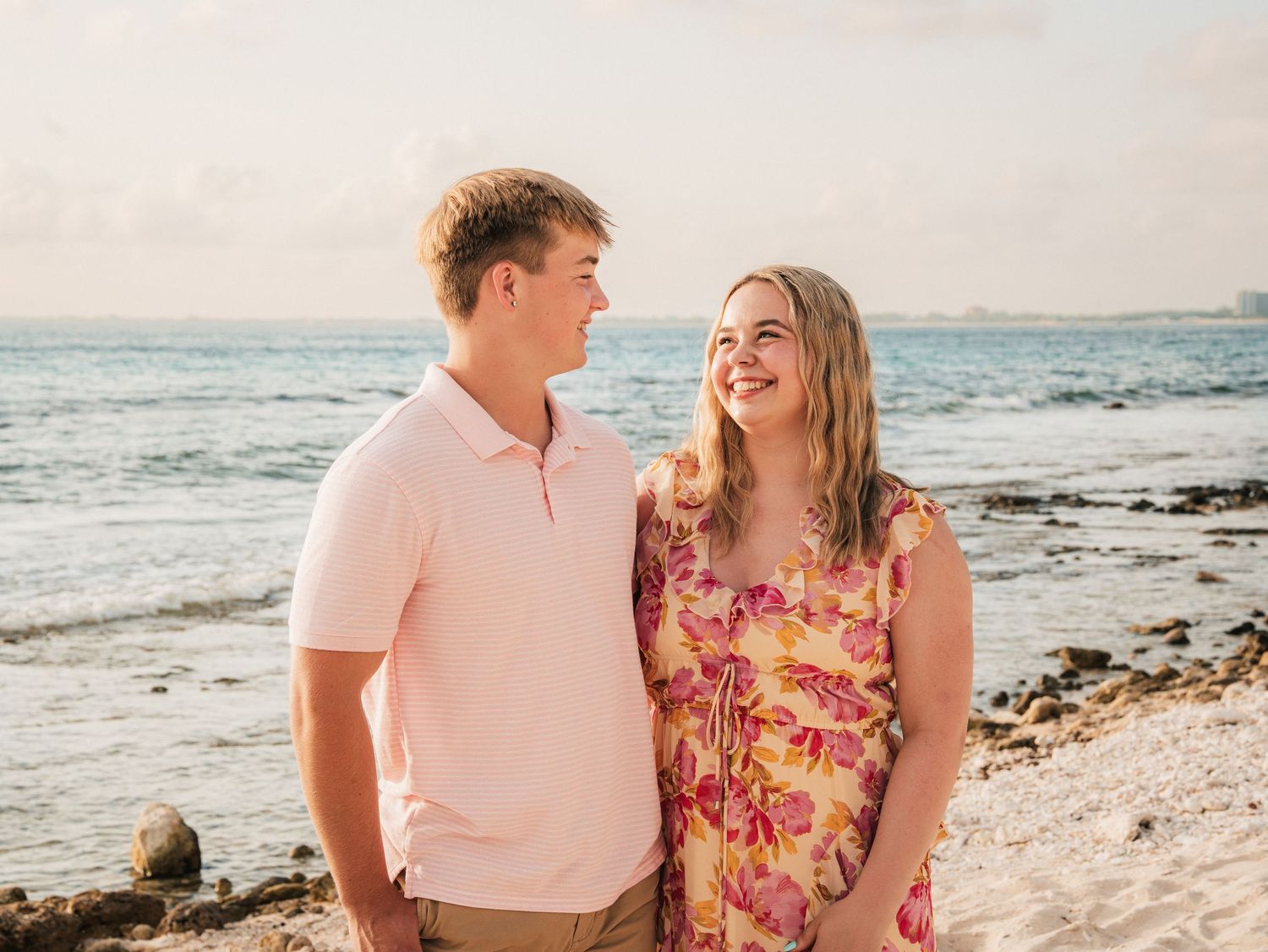 A smiling couple shares a tender moment on a sandy beach, the woman in a floral dress and man in pink.