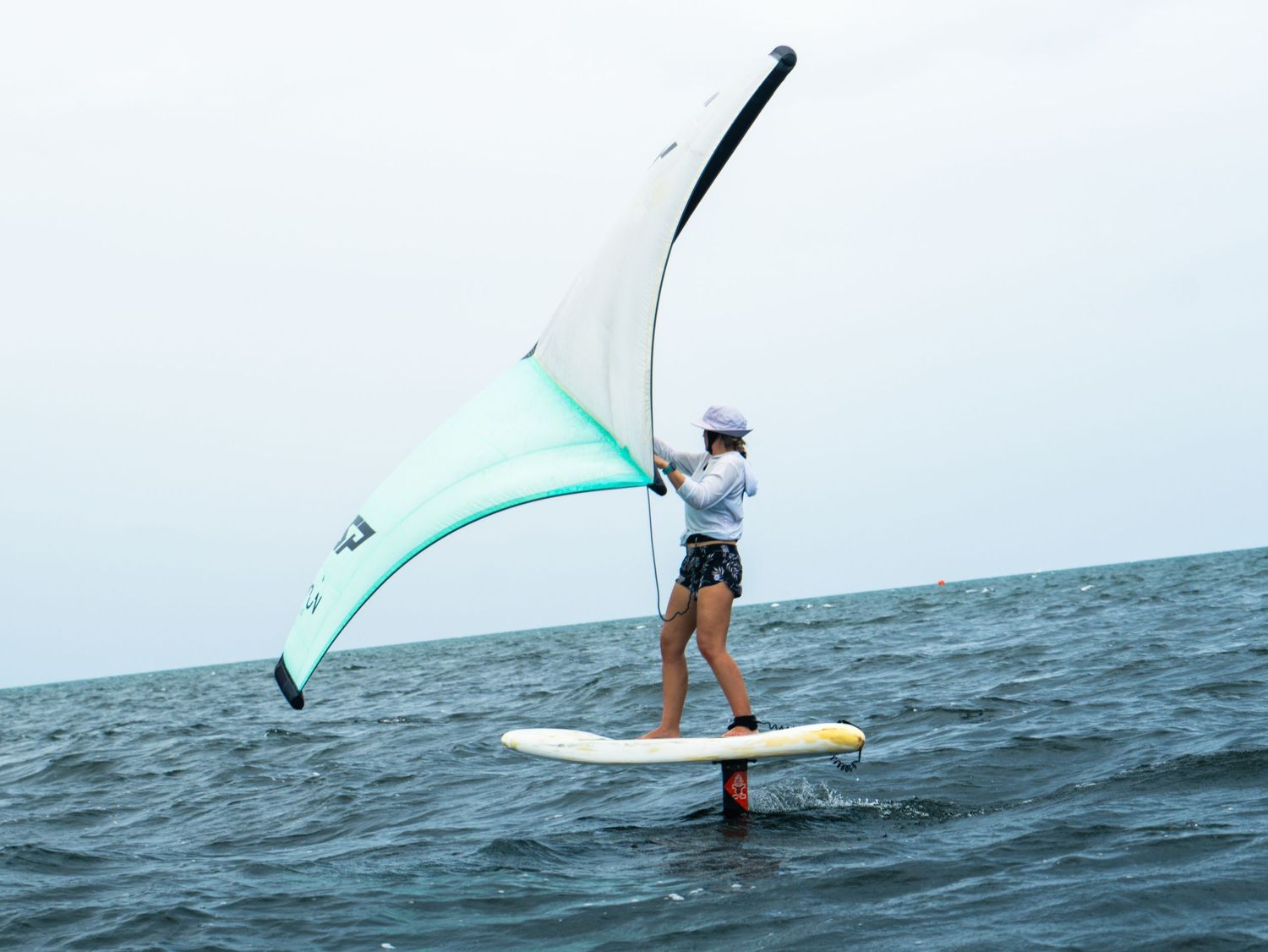 Woman wing foiling on ocean water, balancing on hydrofoil board while holding inflatable wing sail.