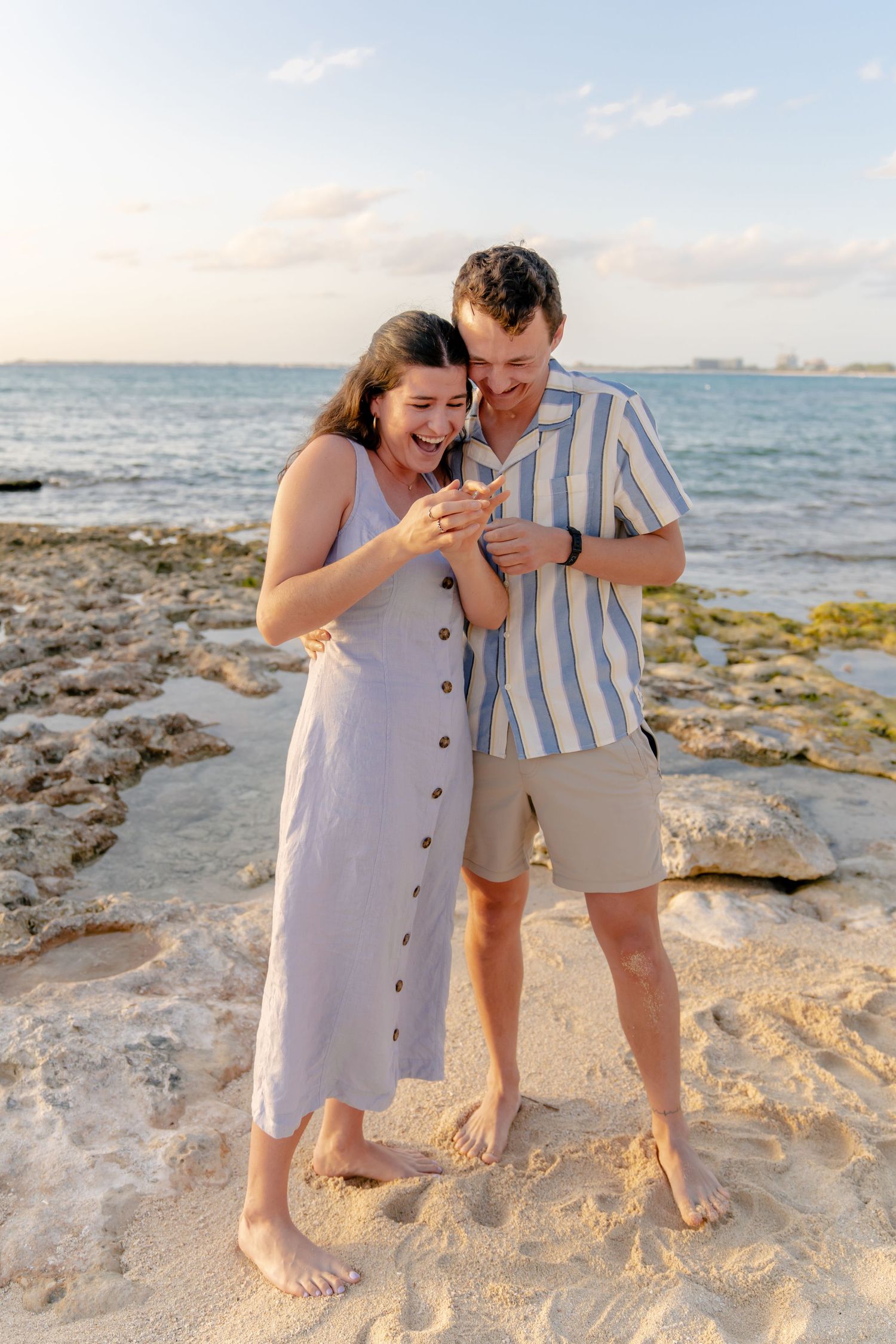 A couple shares a joyful moment looking at their phones together on a beach at sunset.
