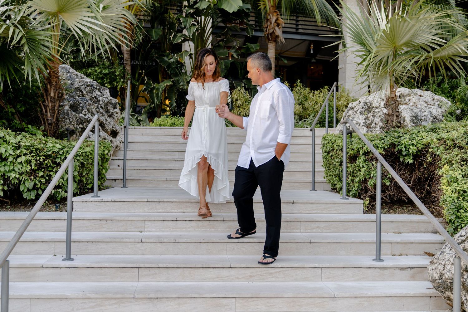 A couple descends white stone steps surrounded by lush tropical landscaping.