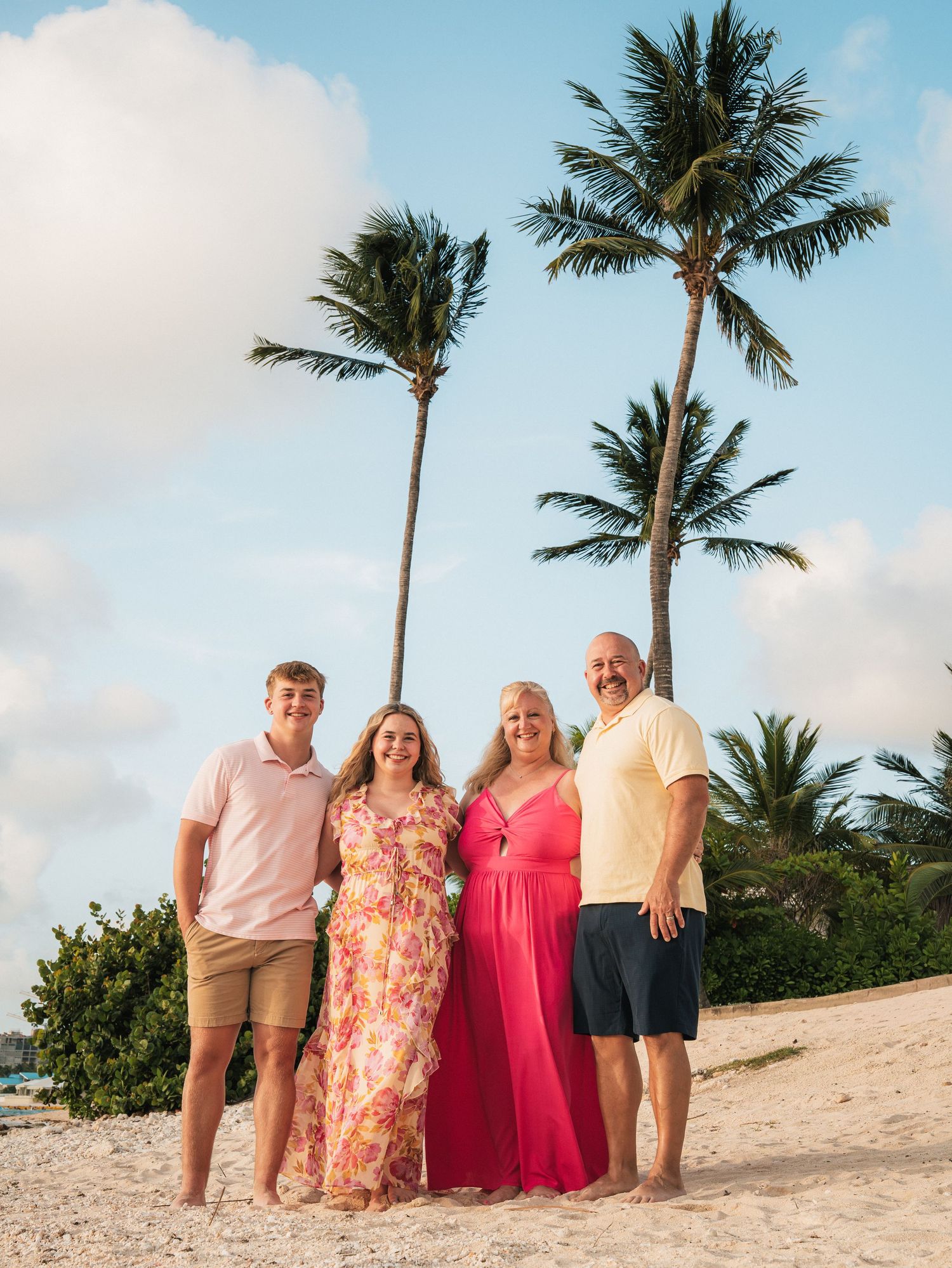 Family of four smiling on a sandy beach with palm trees and blue sky in a tropical destination.