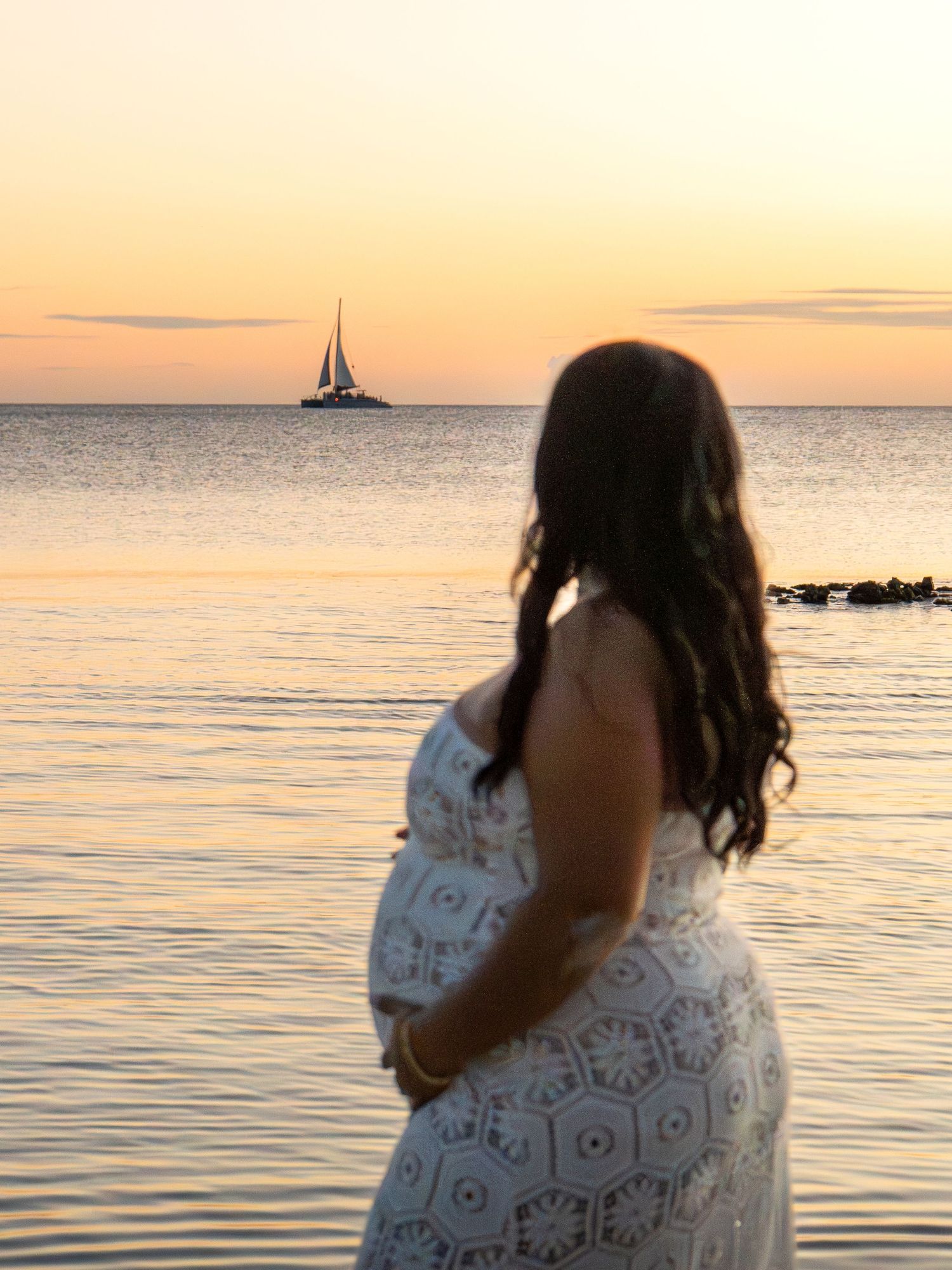 Pregnant woman in white lace dress gazing at sailboat on calm ocean at sunset.