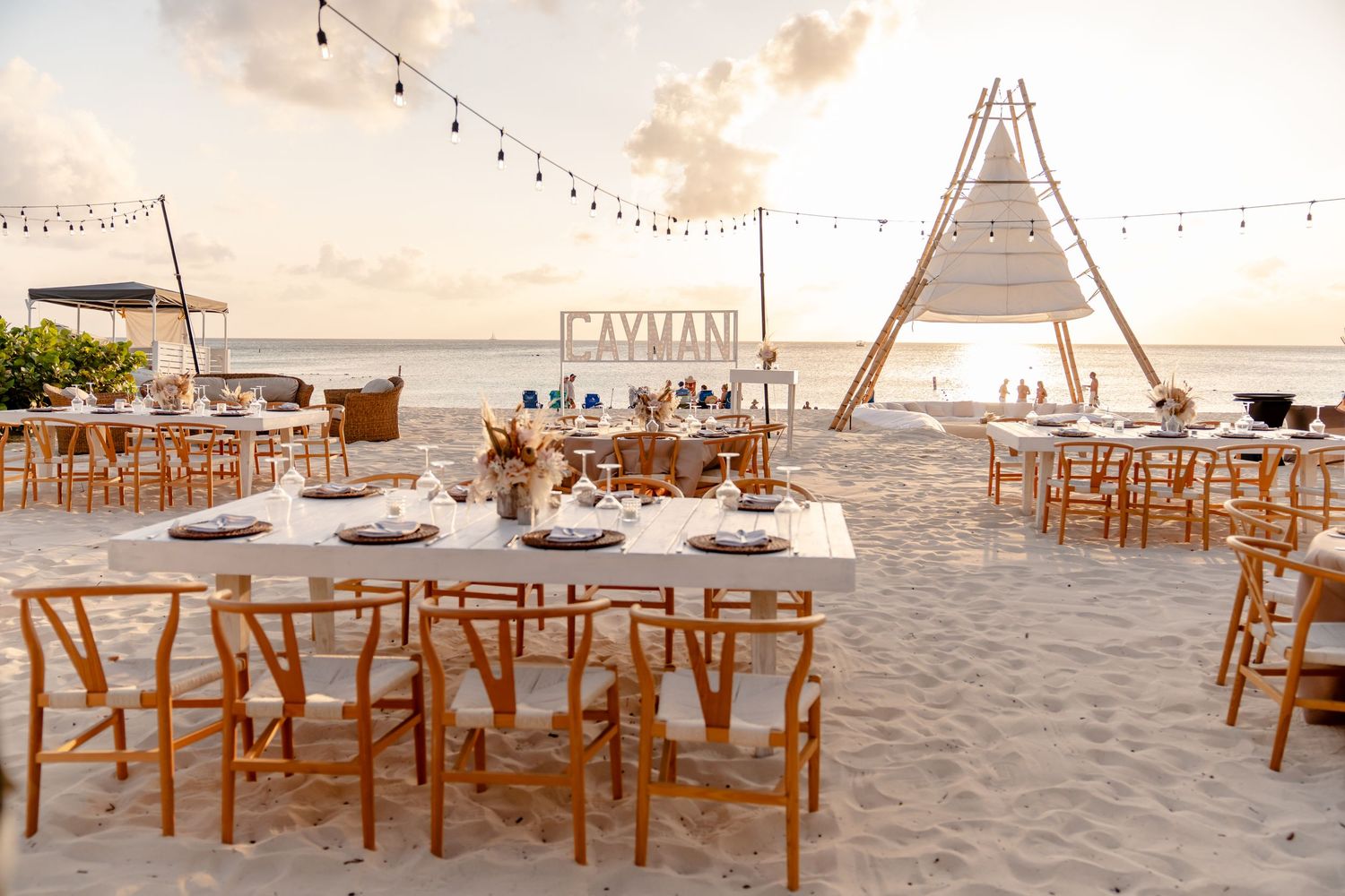 Beach wedding reception setup with wooden chairs and string lights at sunset.