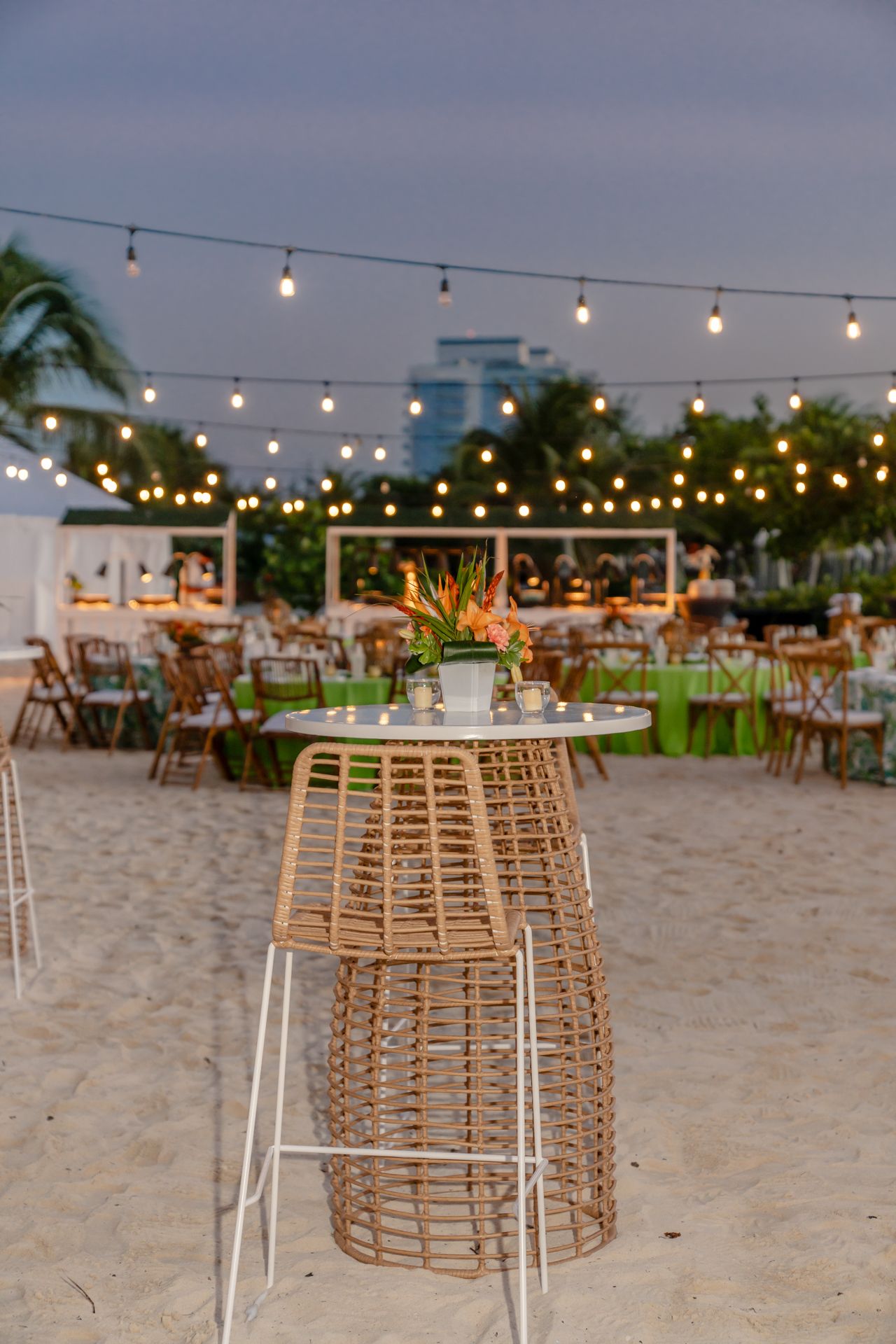 Beach wedding reception setup with rattan furniture and string lights on sandy shore at dusk.