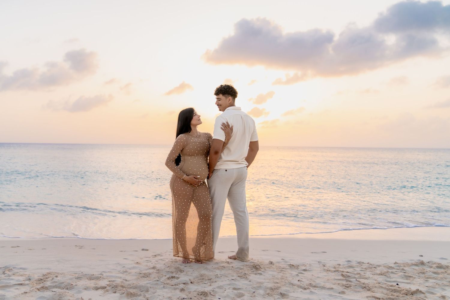 Beach maternity photoshoot featuring couple in white outfits against sunset ocean backdrop.