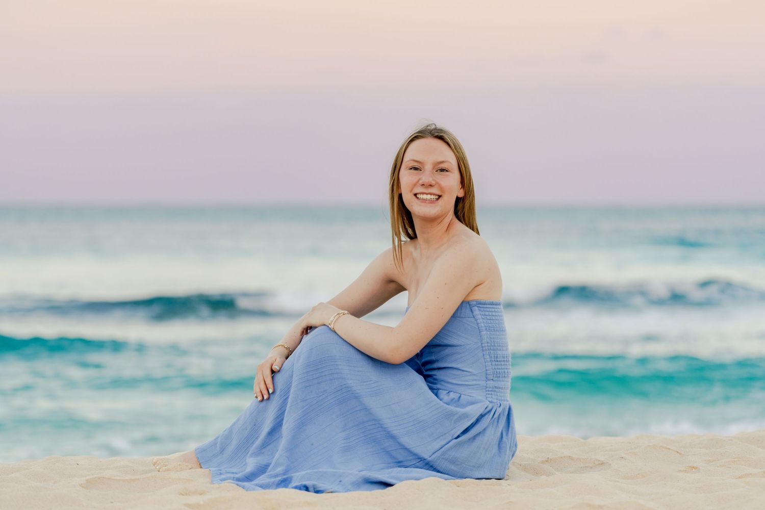 Person in blue dress sitting on sandy beach during sunset with waves in background.