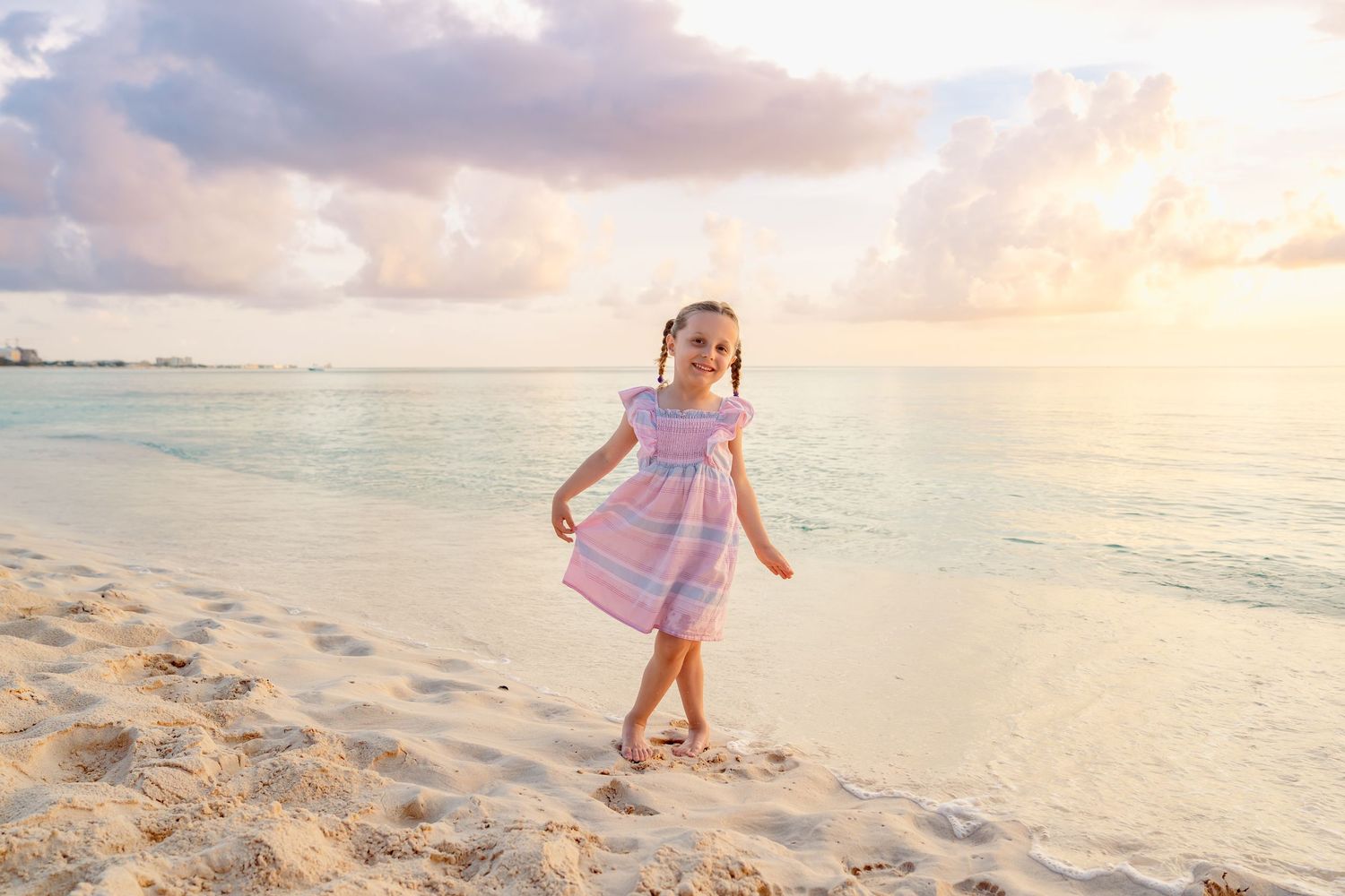 A happy little girl in a pink dress plays on a sandy beach at sunset with turquoise ocean waves behind her.