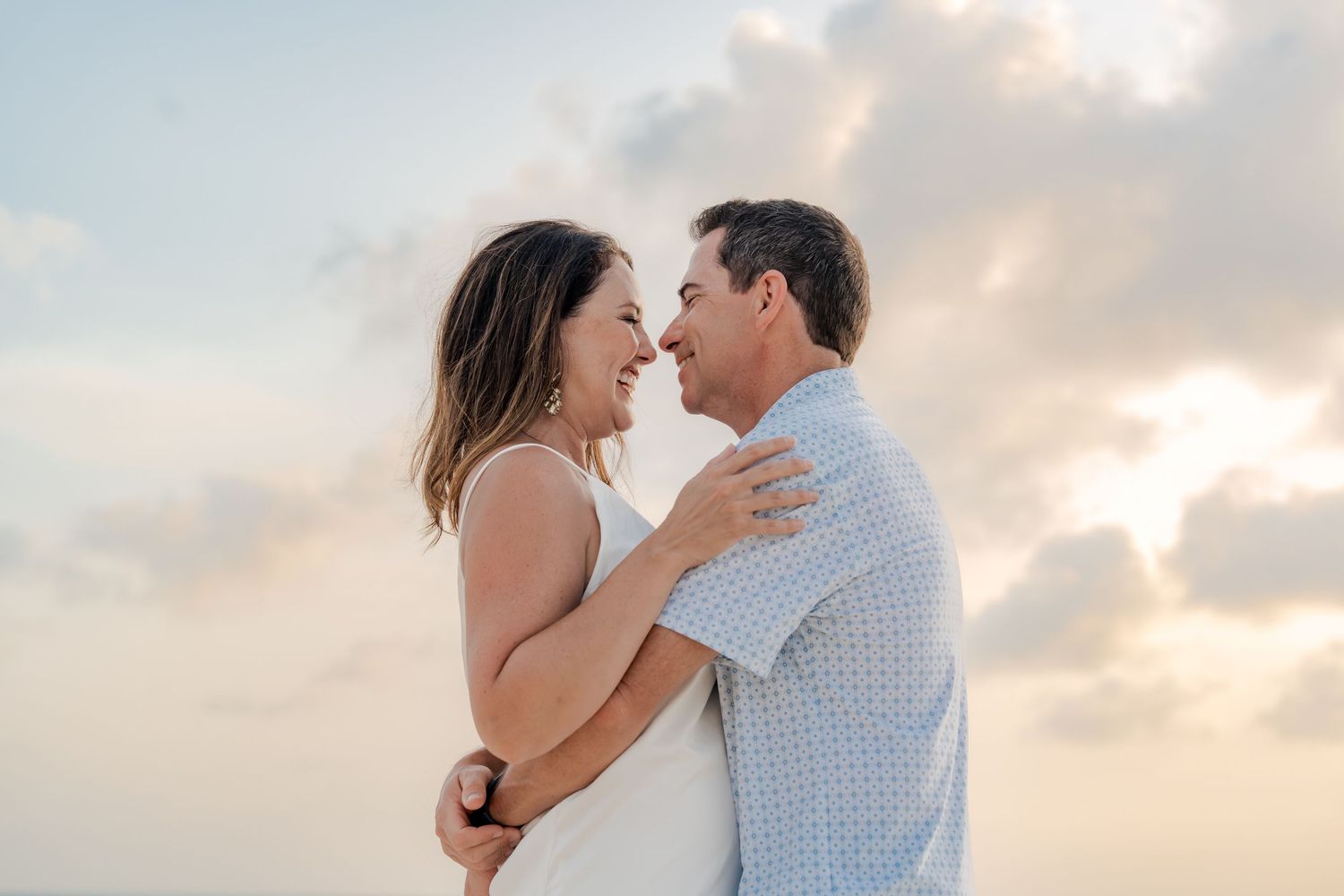 A couple embraces and smiles at each other against a soft cloudy sky at sunset on a beach.