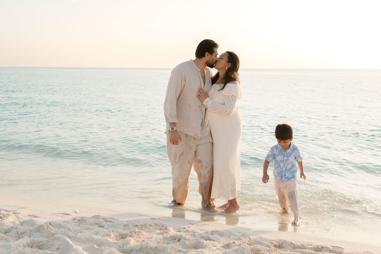 Couple kissing on a sandy beach at sunset while their young child plays nearby in the shallow ocean water.
