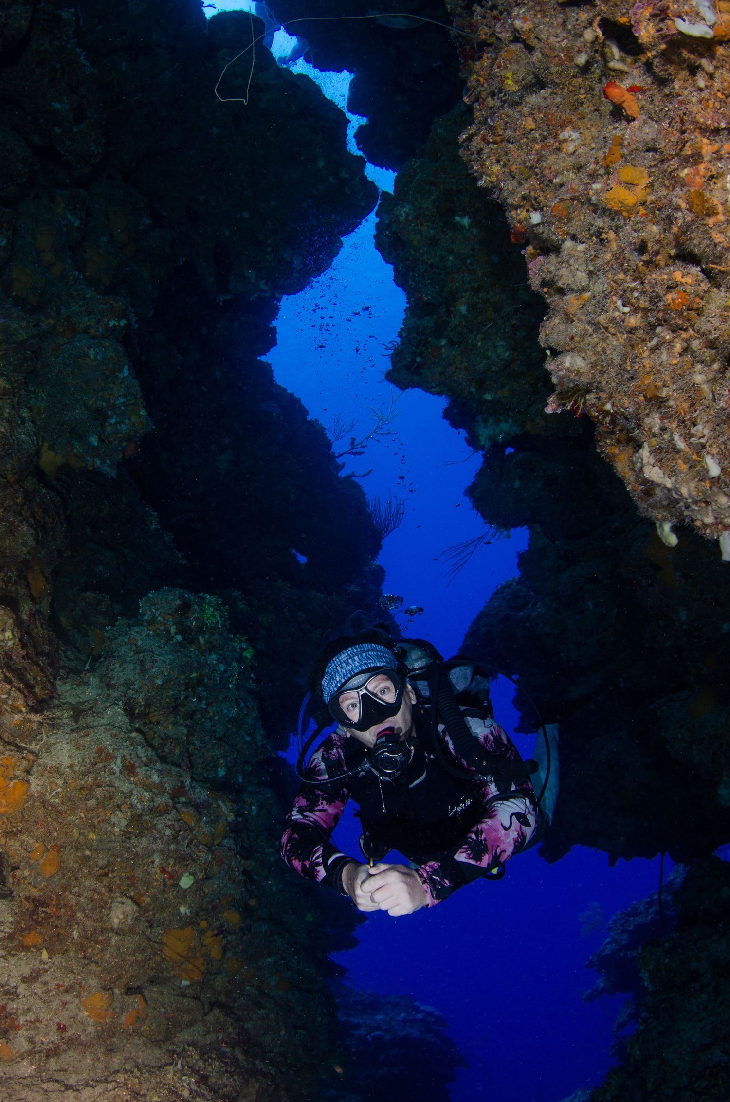 Scuba divers exploring dark underwater cave with bright blue water visible through opening.