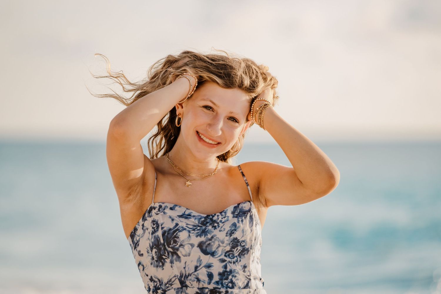 A person in a floral dress poses playfully on a beach with turquoise water and blue sky in the background.