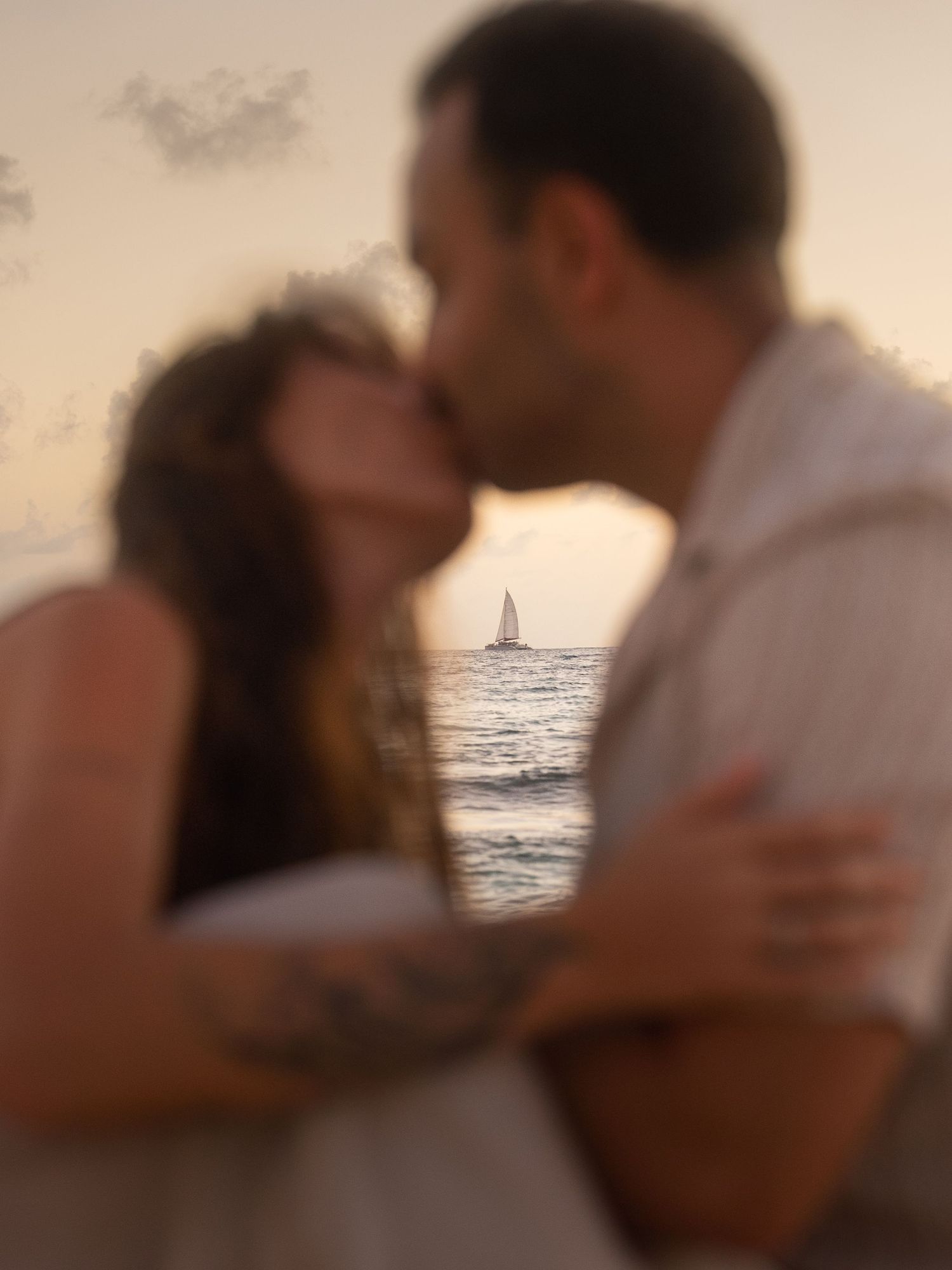 Couple sharing a tender kiss on the beach at sunset with sailboat in background.