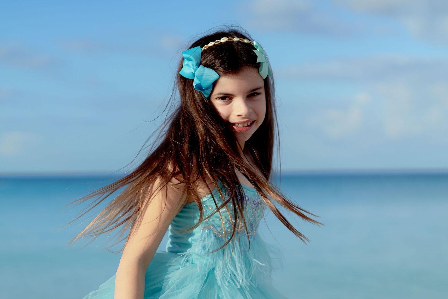 A person in a turquoise dress stands on a beach with wind-swept hair against a bright blue ocean backdrop.