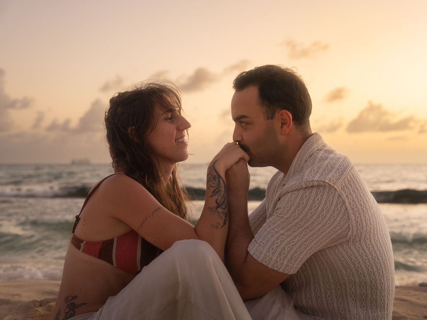 Couple sitting together on a beach at sunset, sharing a tender romantic moment by the sea.
