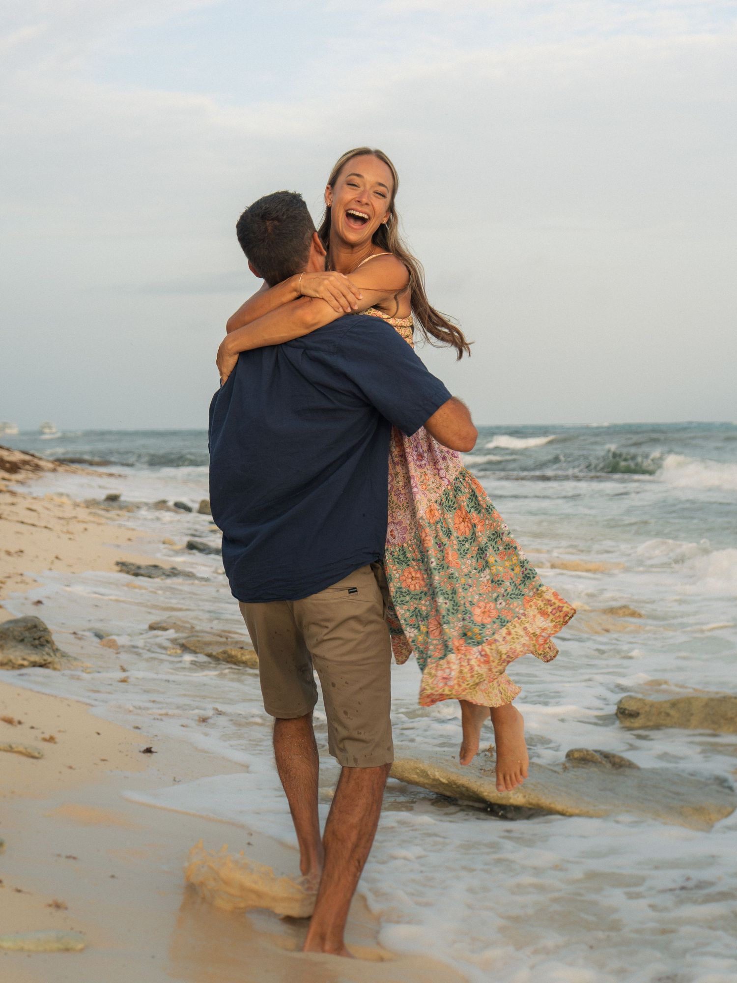Couple embracing and laughing on a sandy beach with ocean waves, woman in floral dress lifted by man in navy shirt.