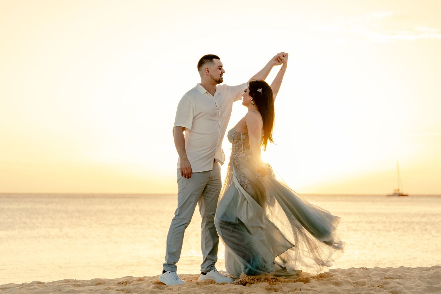 A couple dances romantically on a beach at sunset with flowing dress movement creating an ethereal scene.
