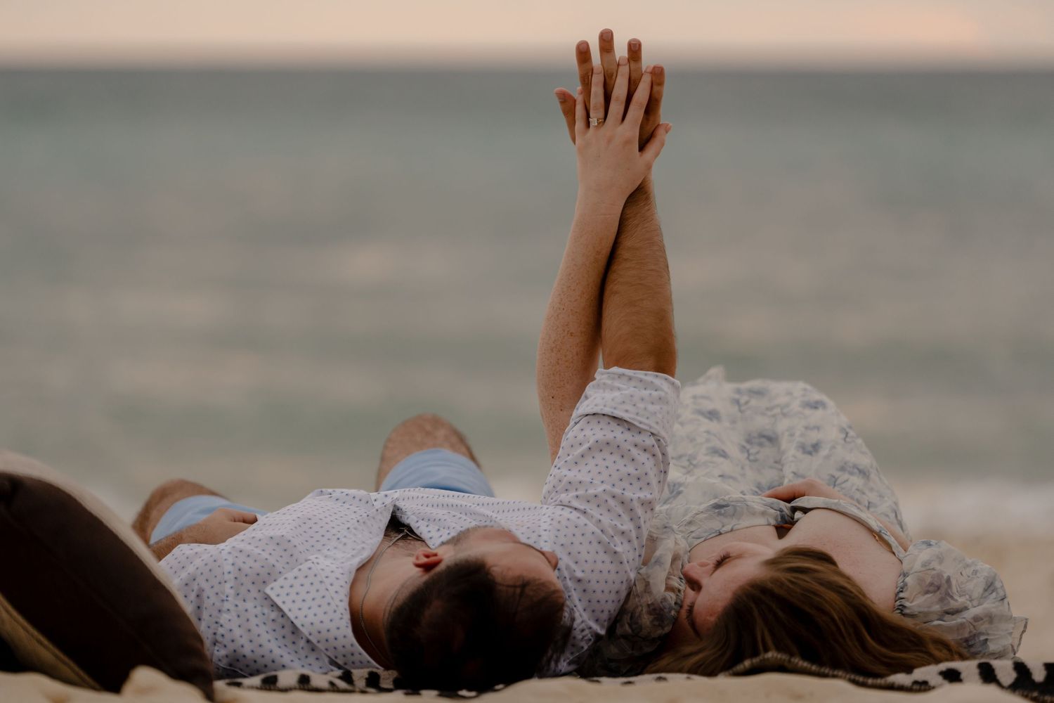 Two people lying on beach sand with raised hands against sunset sky.
