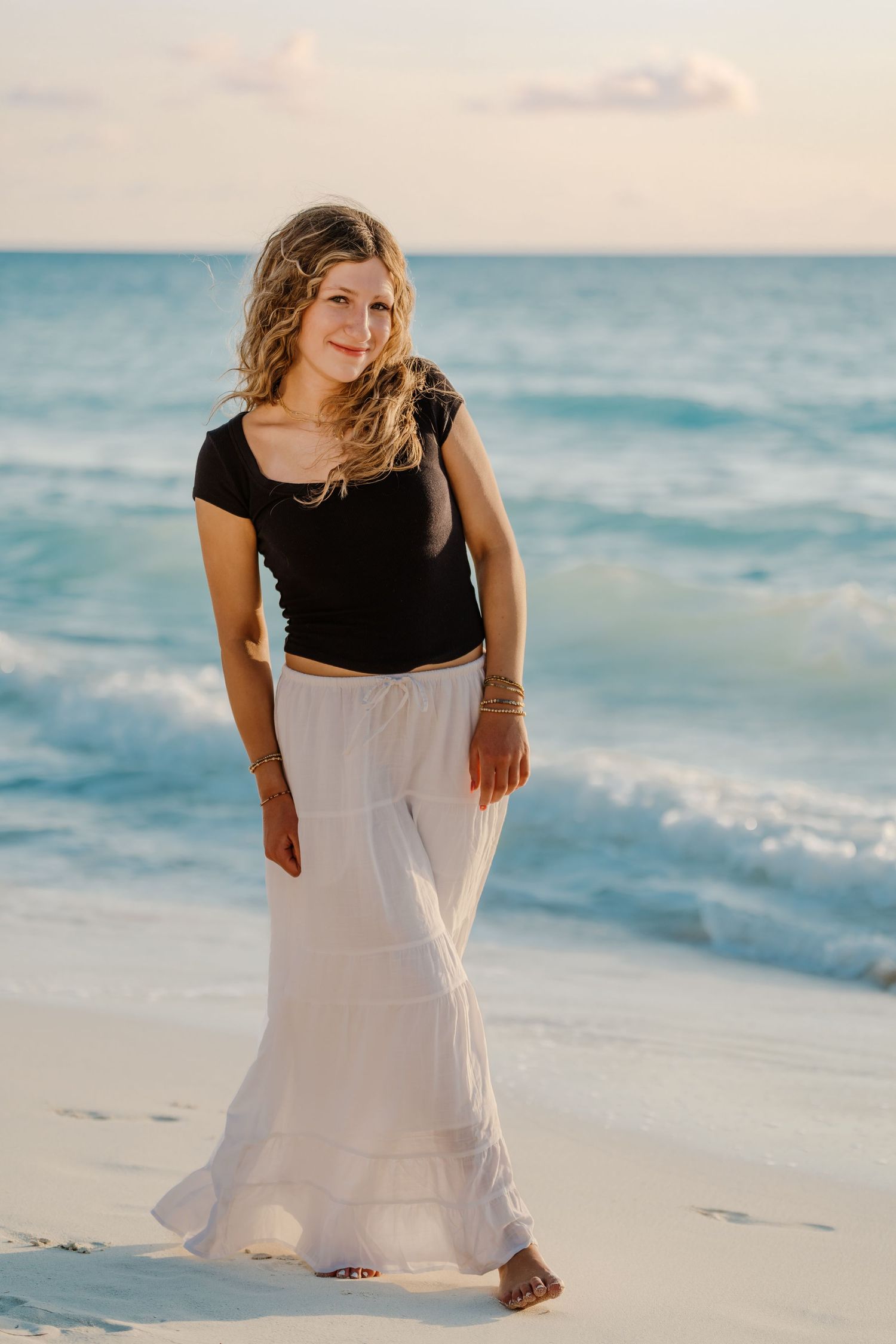 A person in a black top and white skirt stands on a sandy beach with ocean waves in the background.