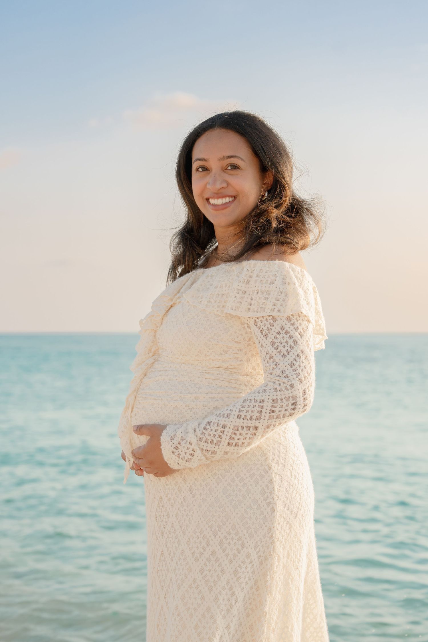 Smiling pregnant woman in white lace dress cradling baby bump on a serene beach with turquoise ocean backdrop.
