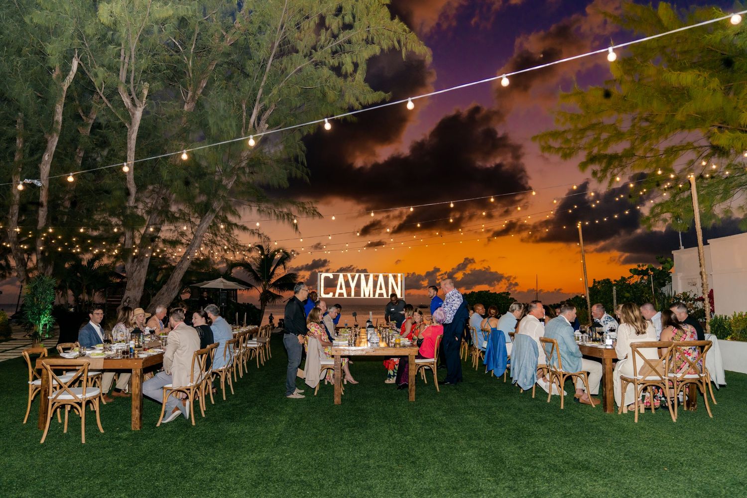 String lights illuminate an outdoor beach dining setup with tables and chairs at dusk.