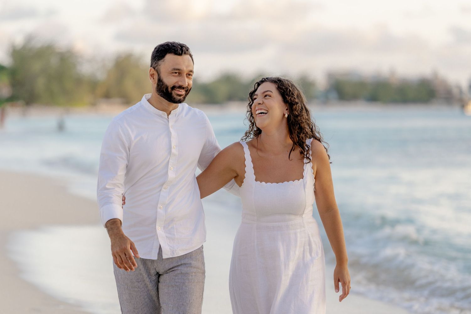 Couple in white and grey attire walking together on a sandy beach.
