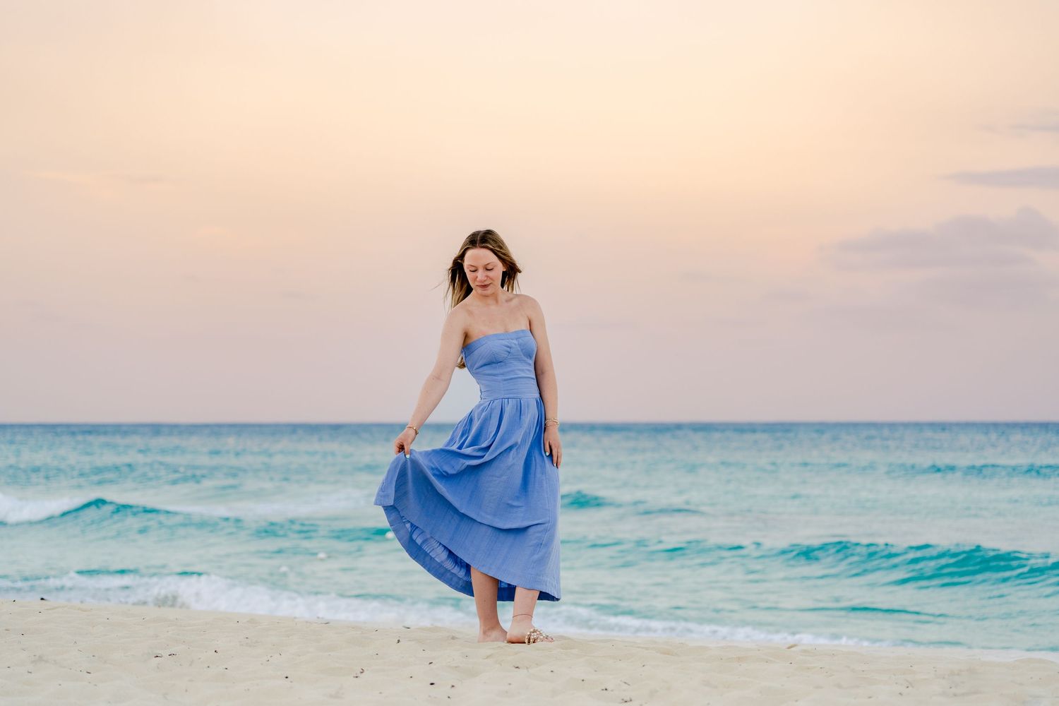 Woman in flowing blue dress walking along beach at sunset with turquoise ocean waves in background.