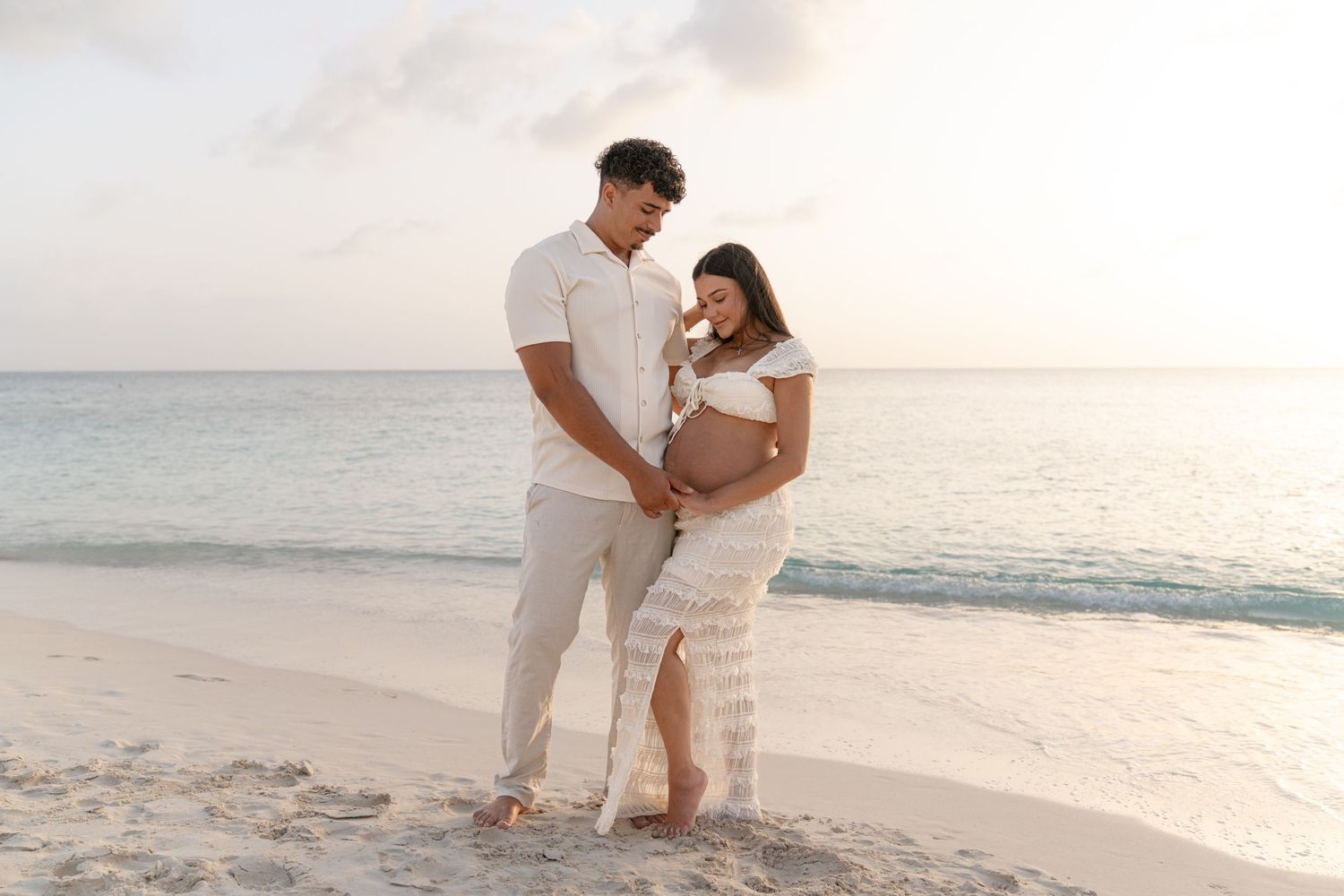 Couple in white outfits embracing on a beach at sunset with ocean waves in the background.