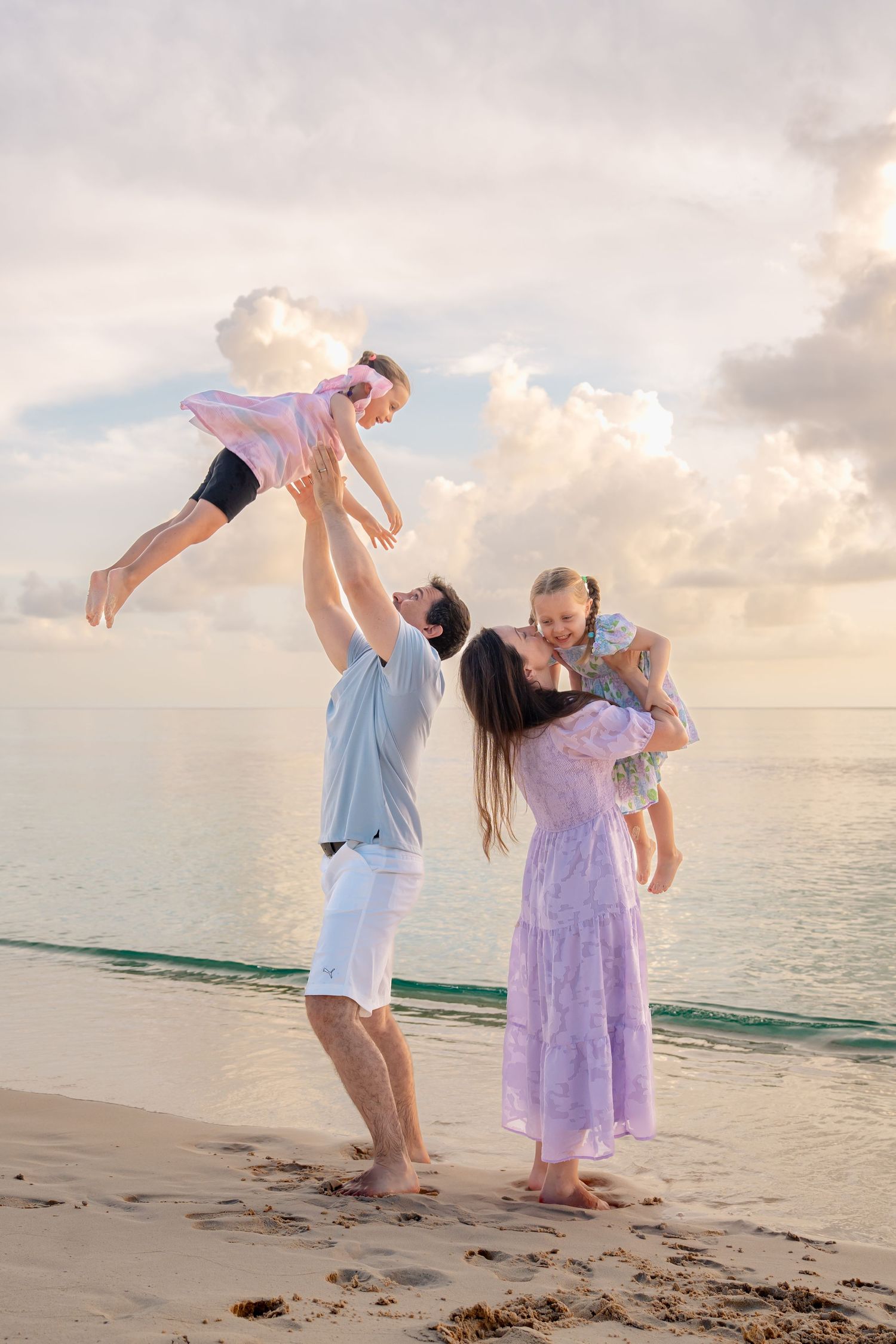 Family of four enjoying a beach vacation at sunset, parents playfully lifting two young children on a tropical sandy shore.