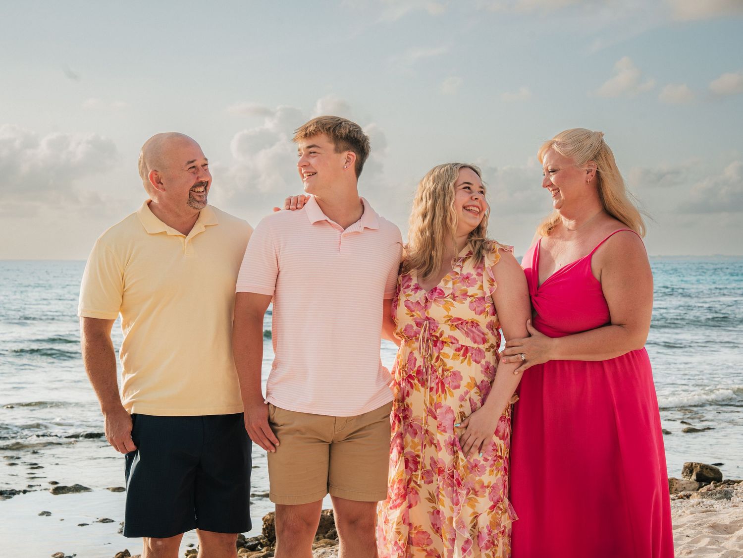 Family of four smiling together on a tropical beach, wearing colorful summer outfits with ocean waves in the background.