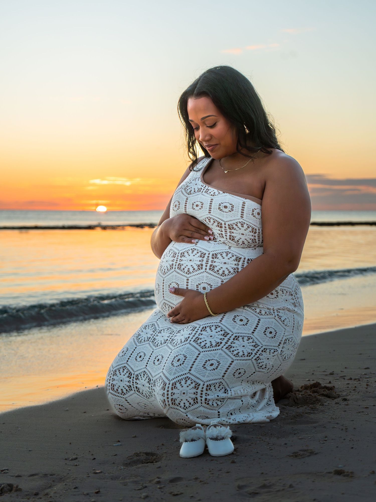 Pregnant woman in white lace dress kneeling on beach at sunset, cradling baby bump with tiny shoes in front.