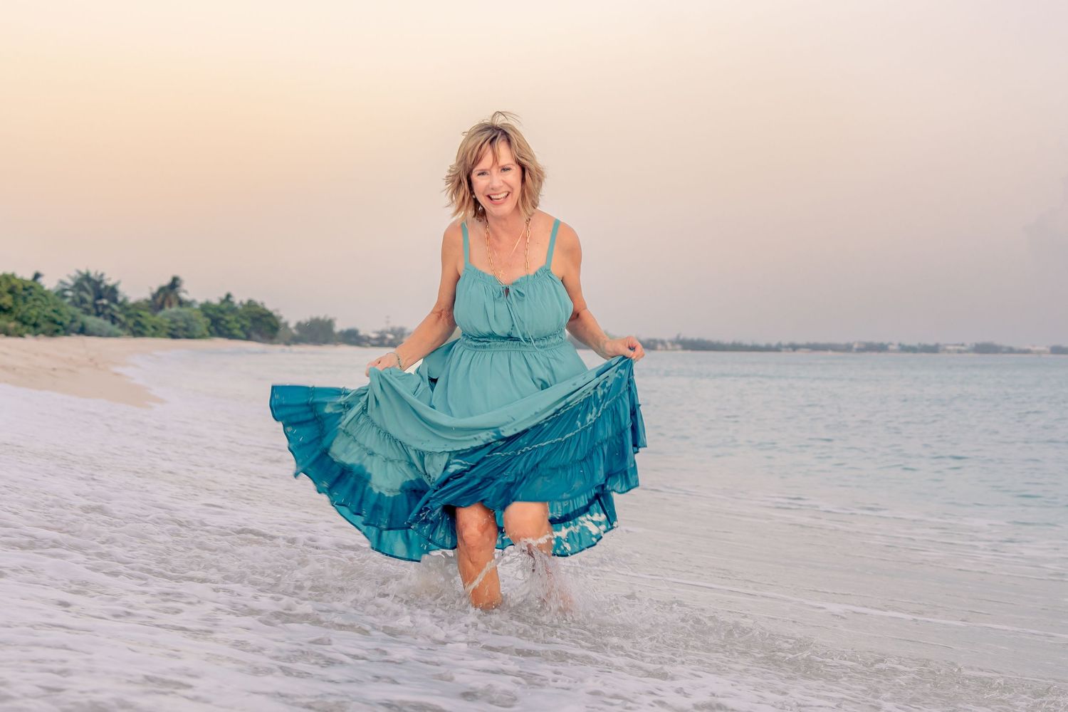 Woman in teal dress joyfully splashing in ocean waves on a sandy beach at sunset.