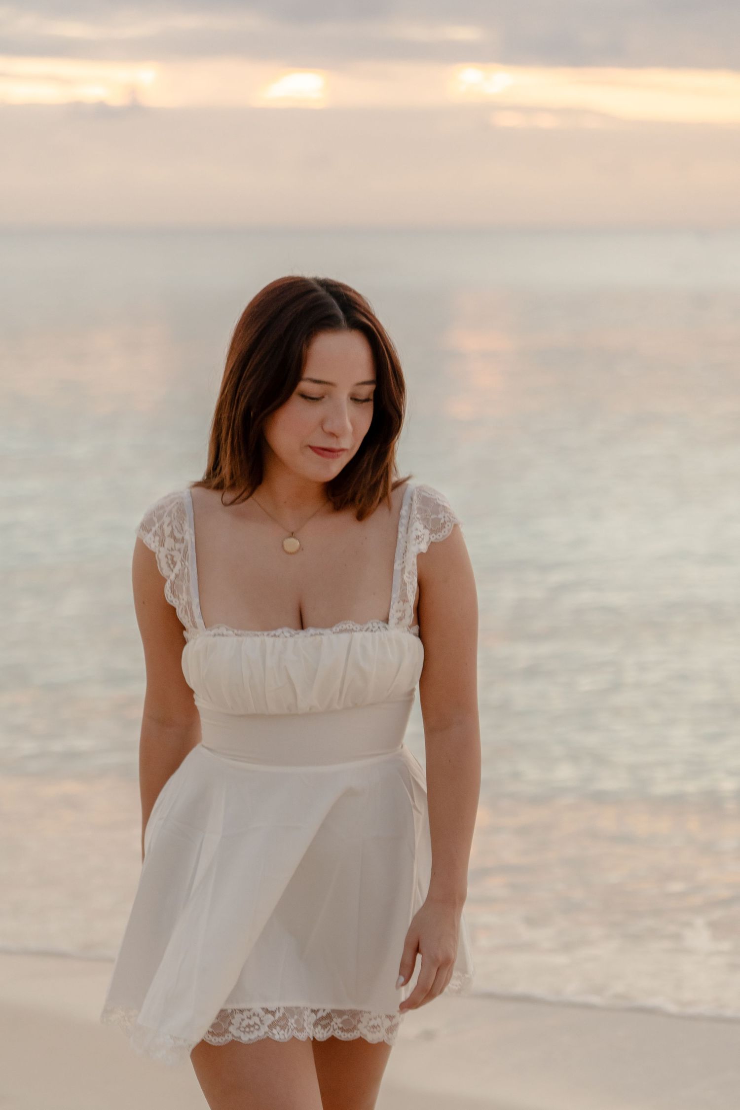 Woman wearing white eyelet dress poses by ocean during sunset.
