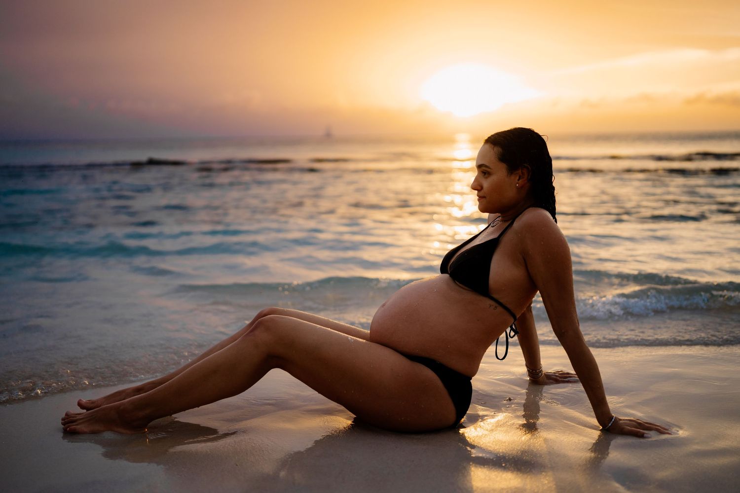 Silhouette of pregnant woman posing on beach at sunset with waves in background.