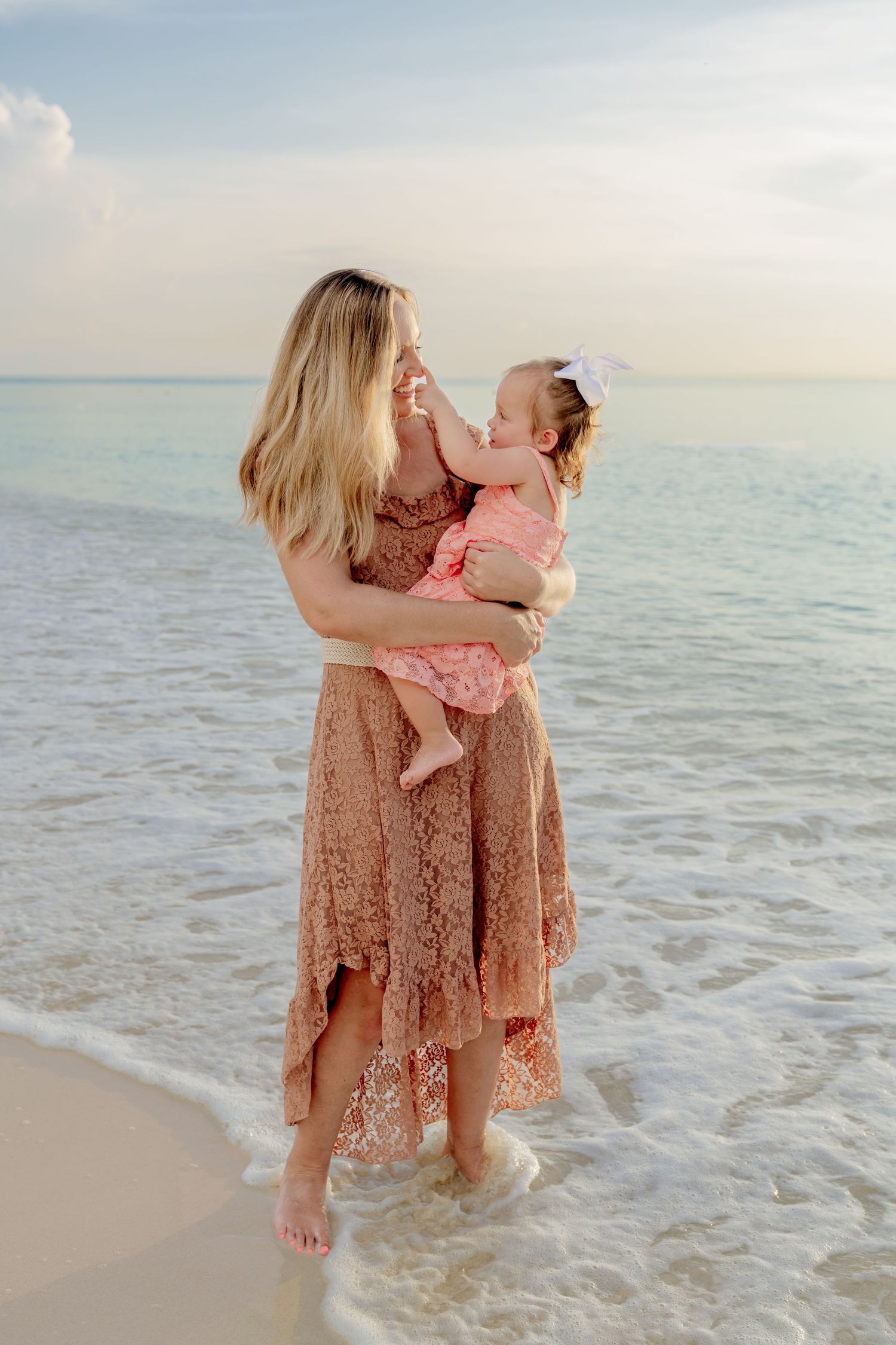 Tender moment between two people in flowing dresses standing in shallow ocean waters at dusk.