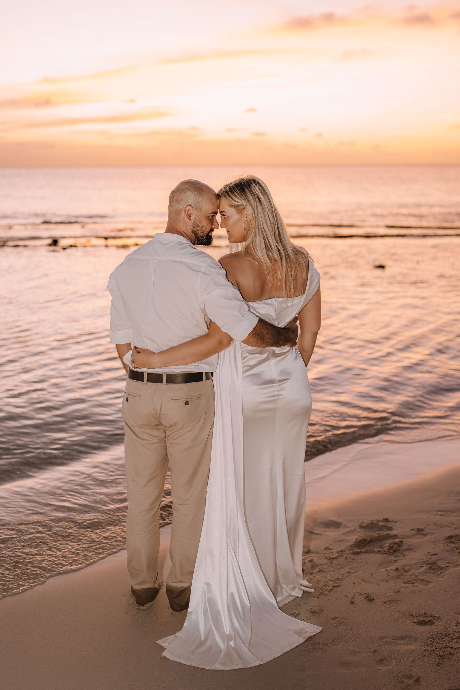 Romantic couple embraces on beach at sunset with flowing white dress and khaki attire.