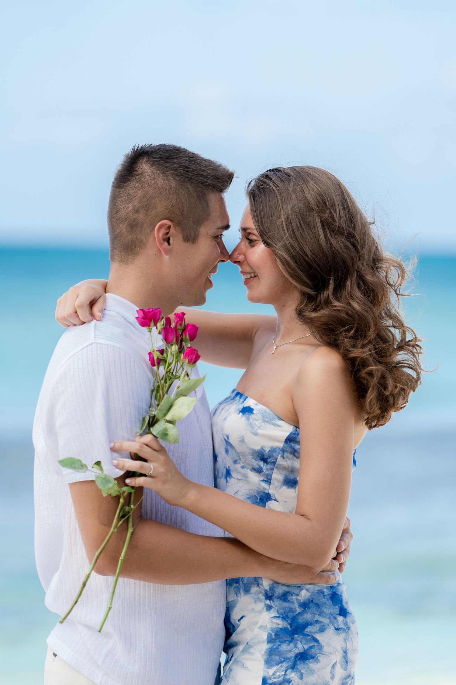 Couple sharing intimate moment with pink flowers on beach with ocean backdrop.